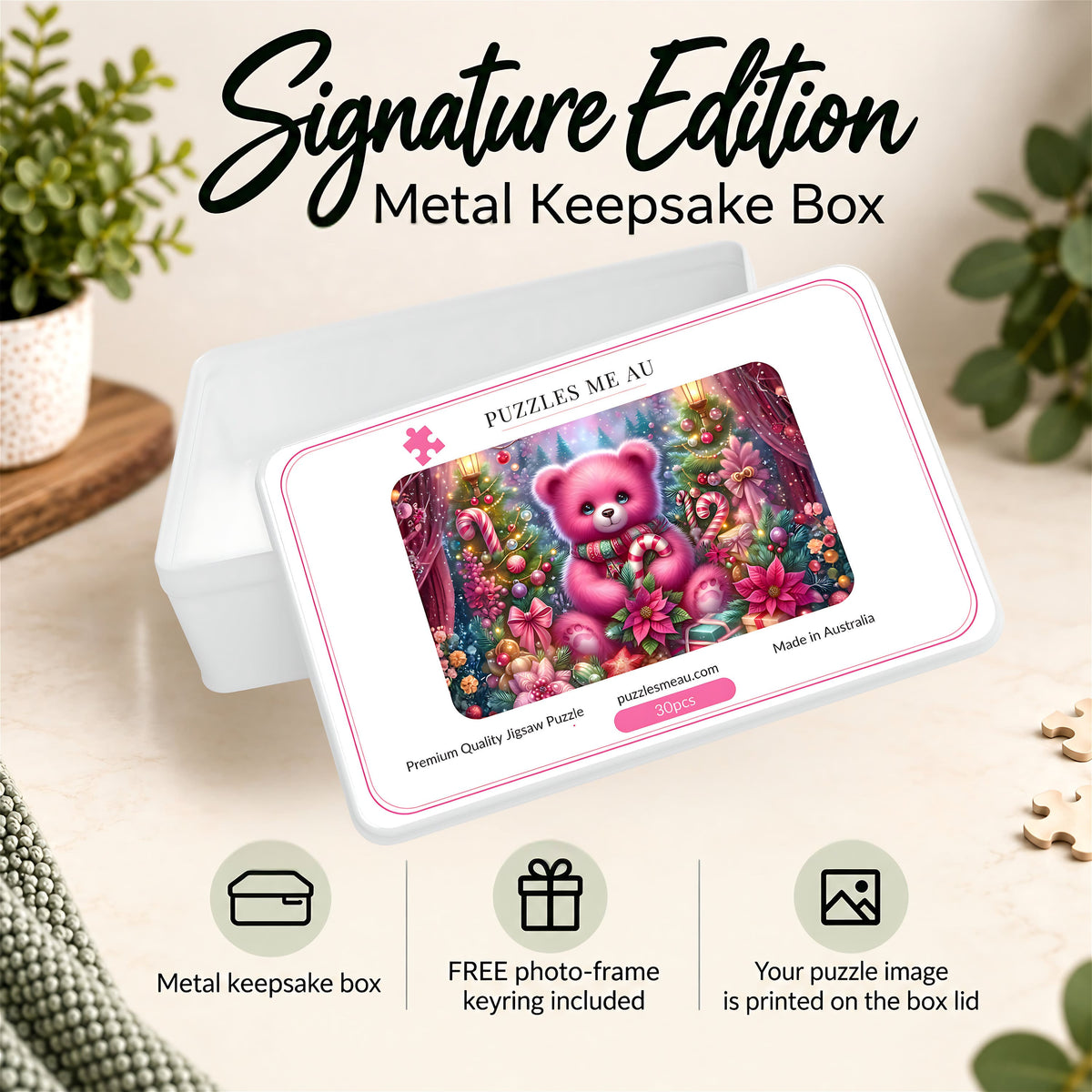A metal keepsake box shown in a styled indoor setting with plants and tabletop decor. The box lid features a colorful printed illustration of a pink teddy bear in a floral scene, with the text 'PUZZLES ME AU' and 'Made in Australia.