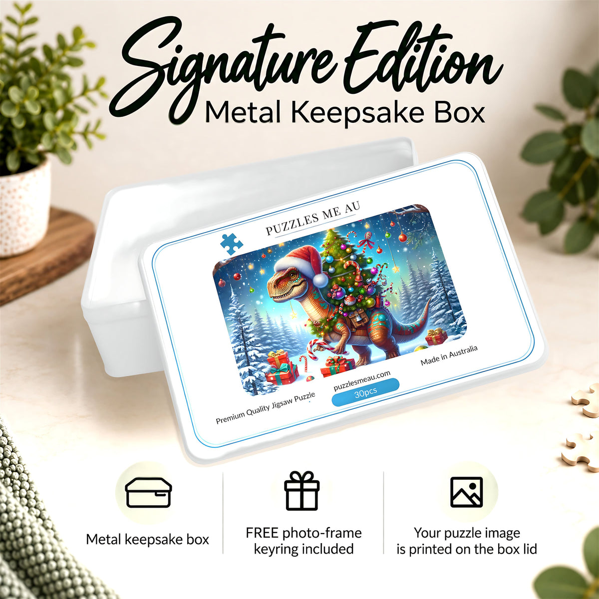 A metal keepsake box shown in a styled indoor setting with plants and decorative objects. The box lid features a printed holiday dinosaur puzzle design with text including 'Puzzles Me Au' and 'Signature Edition Metal Keepsake Box.