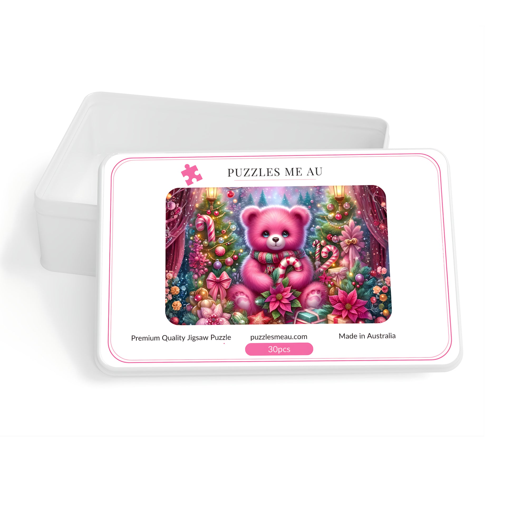 A jigsaw puzzle box with a white lid leaning behind it on a white background. The front of the box shows a colorful illustration of a pink teddy bear in a decorated Christmas tree scene, with the text 'PUZZLES ME AU' and 'puzzlesmeau.